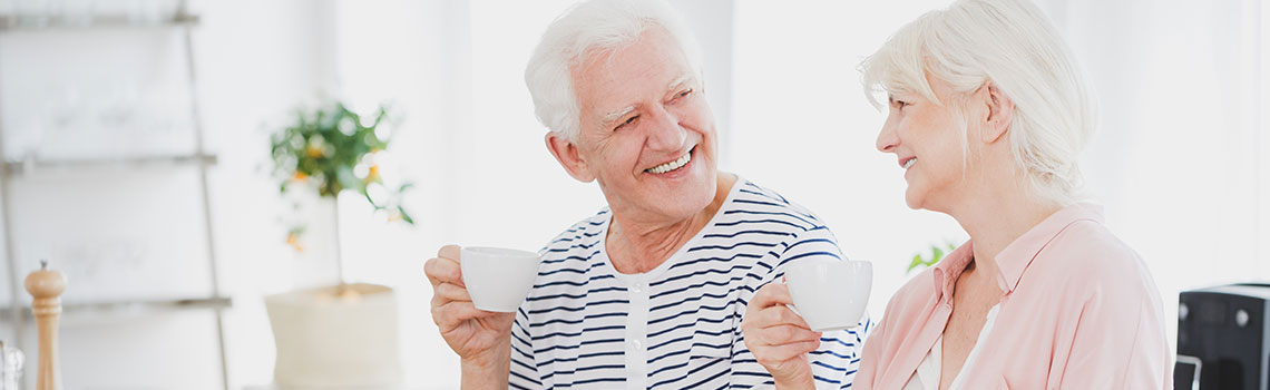 older couple sitting in their home smiling at each other and enjoying coffee in white mugs